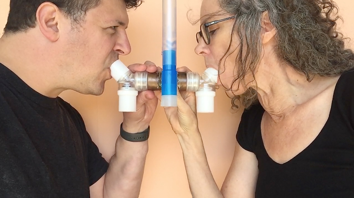 Two people face each other side-on, gripping a medical breathing tube and inhaling simultaneously. The image is closely cropped around their faces and chests, both wear black t-shirts and have dark hair.