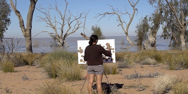 On the banks of an Australian river, a young woman with her back to the viewer, stands painting at an easel.