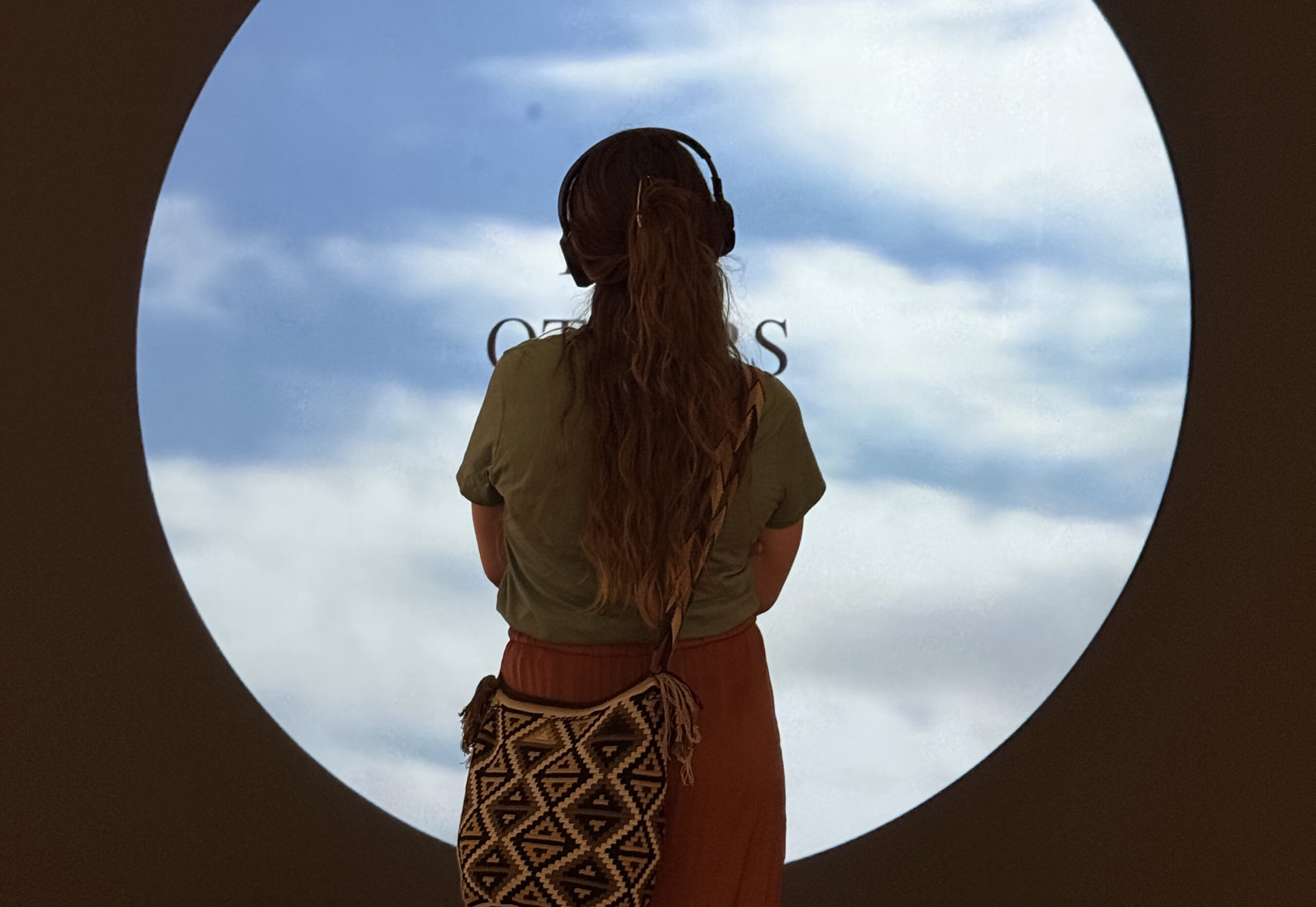Back view of a person with long hair and headphones, looking out through a circular window at a blue sky with clouds.