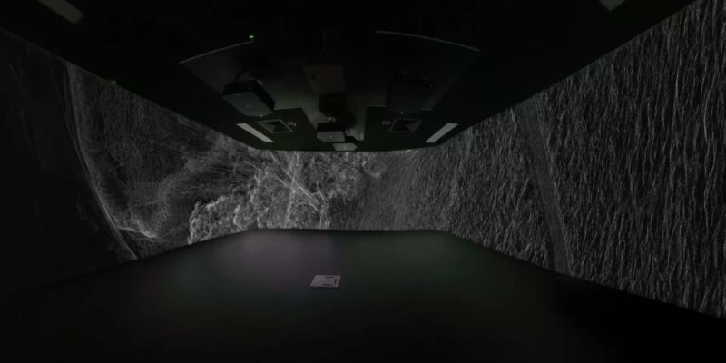 Dim chamber with rough rock walls and a dark panelled ceiling; a small green indicator light and a lone square tile on the floor