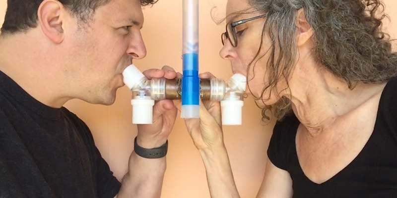 Dr Helen Pynor and Dr Jimmy Breen, documentation, testing the collection of a shared breath sample using the R-tube device. Exhaled DNA will later be Two people face each other side-on, gripping a medical breathing tube and inhaling simultaneously. The image is closely cropped around their faces and chests, both wear black t-shirts and have dark hair.