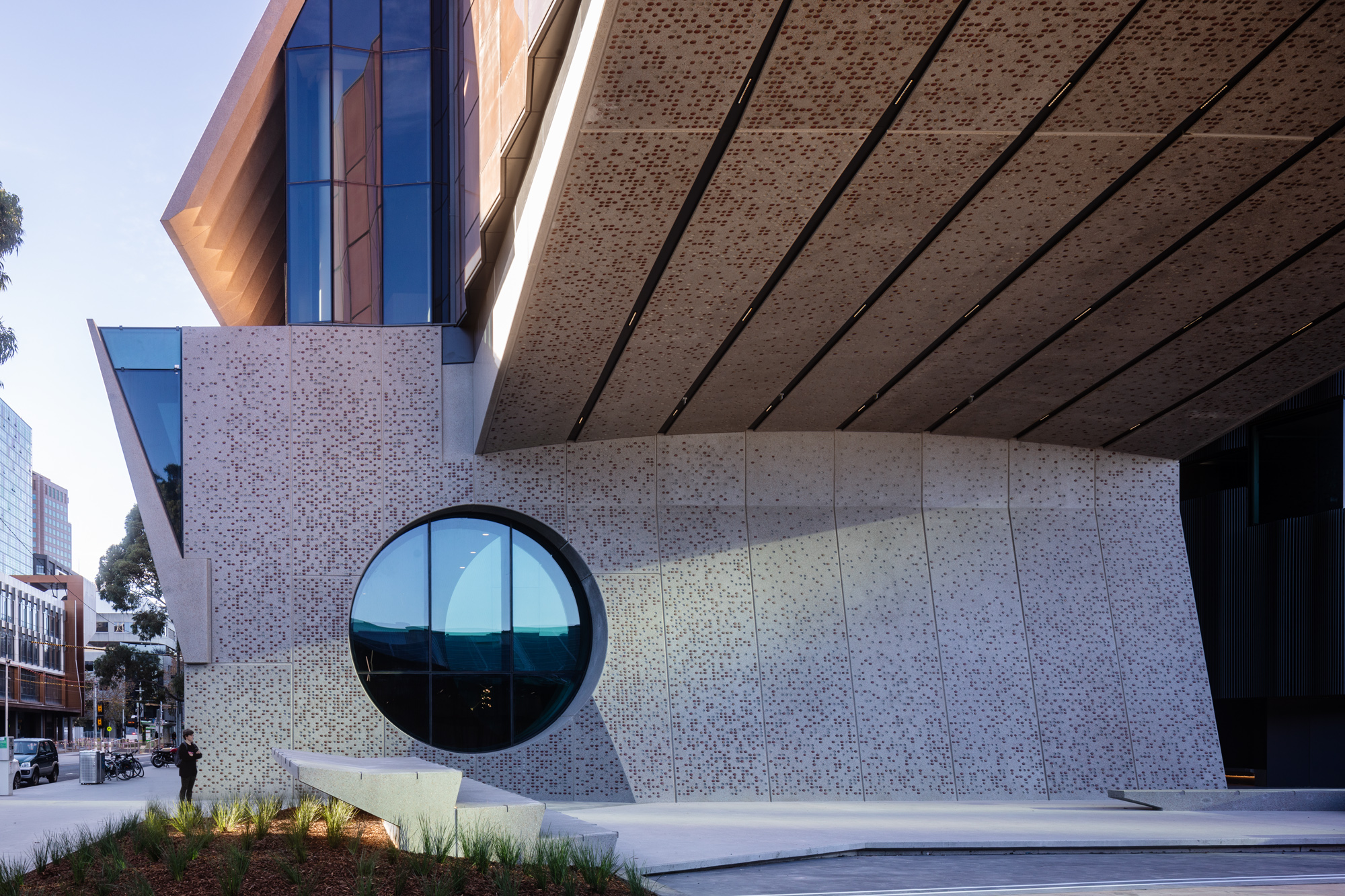 Exterior building with angled design, grey cement walls and red white dots. A roof overhangs a wall with a large, circular window. A small, grassy garden is in the foreground. Blue skies and a street view border the left side.