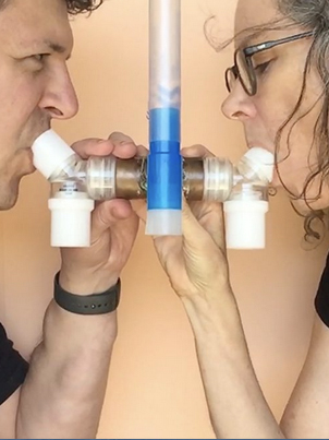 Two people face each other side-on, gripping a medical breathing tube and inhaling simultaneously. The image is closely cropped around their faces and chests, both wear black t-shirts and have dark hair.