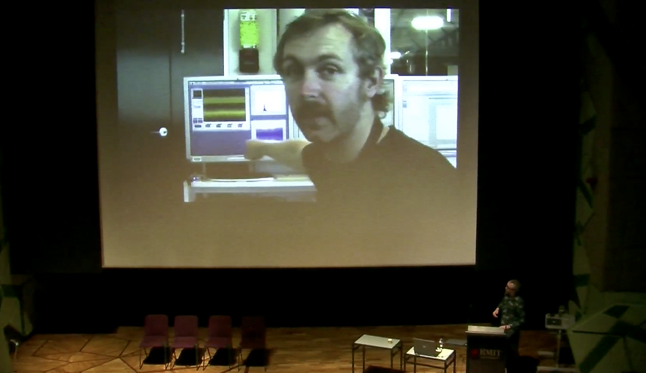 Chris Henschke presenting at the Colliding Ideas symposium, a large screen is projected behind the presenter.