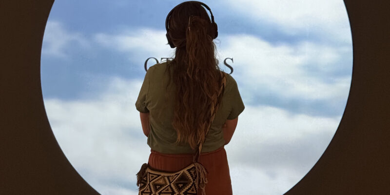 Back view of a person with long hair and headphones, looking out through a circular window at a blue sky with clouds.