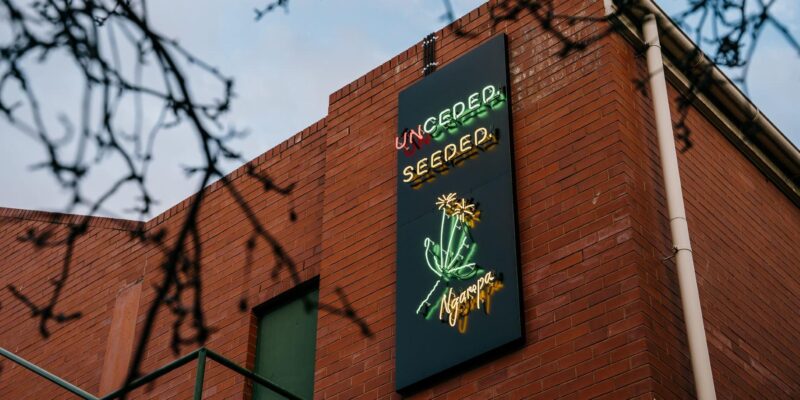 Exterior brick building with a vertical neon sign displaying 'Uncensored Seeded' and a glowing plant graphic on a dark panel.