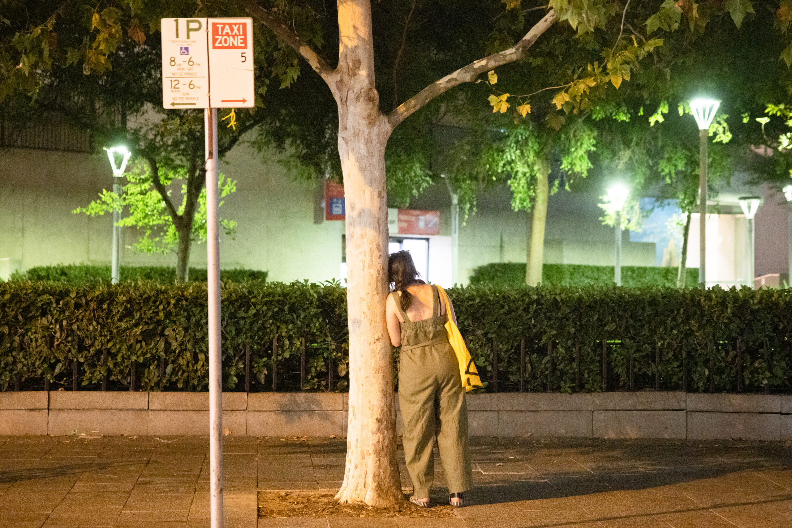 Nighttime street scene: a person in olive overalls stands with a yellow bag, facing away, beside a tree on a sidewalk with greenery in the background.