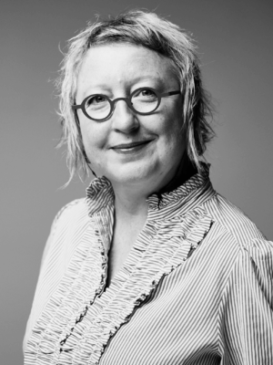Smiling woman with short, light hair and round glasses wearing a striped blouse with ruffles (black and white).