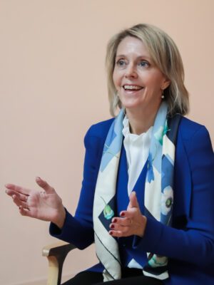 Smiling woman in a blue blazer with a light scarf, gesturing with her hands as she talks in front of a peach wall.