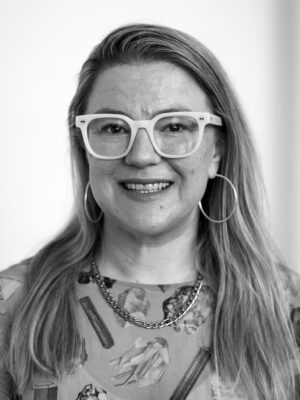 Close-up black-and-white portrait of a smiling woman wearing large white glasses and hoop earrings.